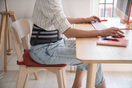 Freelancer man works types on computer keyboard wears back support belt corset on lower back to relieve stress on spine. Treatment of hernia. Back pain health problems, consequences sedentary work. 
