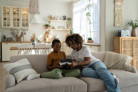 Photo for Black woman mom teaching kid son how to use VR headset for learning and education while sitting on sofa at home. Modern African American family mother and child explore virtual reality together - Royalty Free Image
