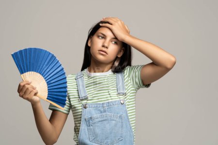 Photo for Too hot. Sweaty tired teenage girl touching forehead using paper fan suffer from heat, feels sluggish. Displeased teen girl cooling in hot summer weather, isolated on studio gray wall. Overheating - Royalty Free Image