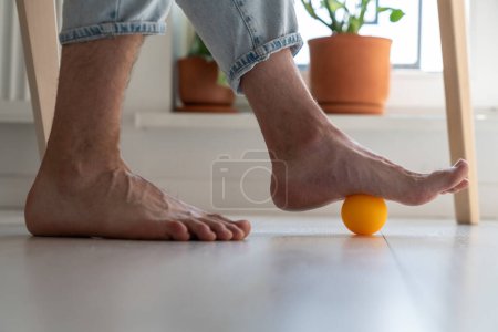 Téléchargez les photos : Homme utilisant la boule de silicone pour le massage des pieds faire de l'exercice physique de prévention pour améliorer la circulation sanguine. Pieds, rétablissement de la colonne vertébrale après un traumatisme hernie, travail sédentaire et concept de style de vie. - en image libre de droit
