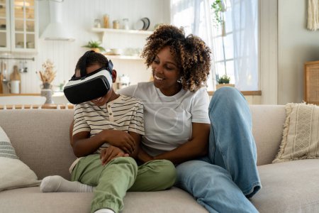 Foto de Feliz alegre familia afroamericana madre e hijo usando auriculares VR mientras se relajan juntos en el sofá, riendo mamá y el niño se divierten mientras juegan juegos de realidad virtual en casa - Imagen libre de derechos