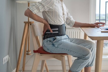 Photo for Man freelancer works types on computer keyboard wearing back support belt corset on lower back during exacerbation to treatment of hernia. Back pain health problems as consequences of sedentary work. - Royalty Free Image