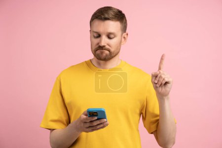 Photo for Sad upset man points index finger up looks in smartphone screen on pink background. Bearded male in yellow t-shirt dissatisfied with reading information in mobile phone. Bad news, problems, troubles. - Royalty Free Image