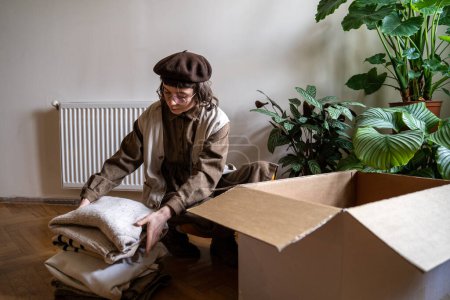 Téléchargez les photos : Une hipster girl éco-activiste empilant de vieux vêtements dans une boîte en carton à la maison, triant les articles indésirables à recycler. Déchets vestimentaires et recyclage, concept de vie consciente. Don, d'occasion, sauver la planète - en image libre de droit