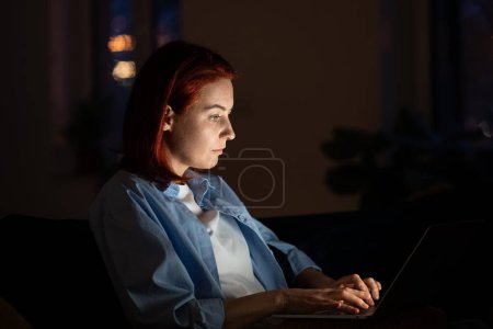 Foto de La mujer programadora enfocada en la habitación oscura se sienta en el trabajo de la silla en el portátil. Workaholic freelancer Mujer europea sentada cómodamente con ordenador en vueltas trabajando en diseño gráfico para app por la noche - Imagen libre de derechos