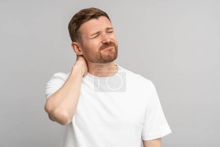 Photo for Portrait of man suffering from cervical osteochondrosis touching rubbing neck. Painful facial expression. Upset male in suffers from pain with closed eyes stands on grey background. Health after 40 - Royalty Free Image
