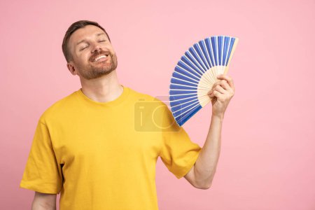 Photo for Smiling relaxed man using paper fan suffer from heat, enjoying fresh air and closed eyes, cools herself due problem no air conditioner at home at summer weather isolated on studio pink background - Royalty Free Image