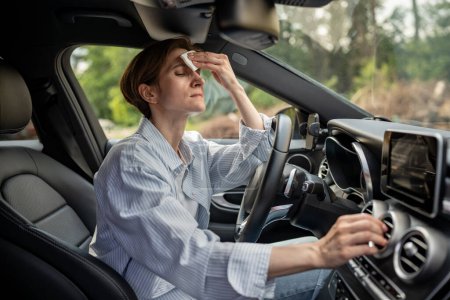 Photo for Woman drives car with broken air conditioning in hot summer weather wipes sweat on forehead with paper napkin. Middle aged female suffering from heat stuffiness. Exhausted overheated stressed driver. - Royalty Free Image