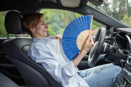 Photo for Tired exhausted middle aged woman waving blue fan suffers from stuffiness driving car on summer hot weather. Overheating, sultriness, high temperature, swelter in car with broken air conditioner. - Royalty Free Image