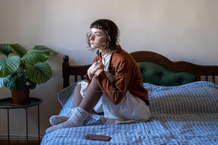Photo for Thoughtful worried teen girl sits on bed sadly look at window waiting call, important message on smartphone. Lonely upset young woman holding knees in hand thinking about problem received bad news - Royalty Free Image