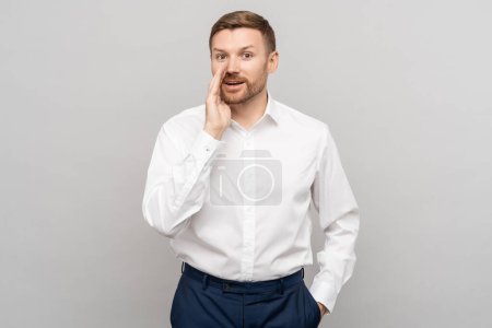Photo for Office worker man whispering with hand near mouth gesture on grey studio background. Advertisement banner. Smiling positive businessman shares a secret offer, looking at camera talking secret. - Royalty Free Image