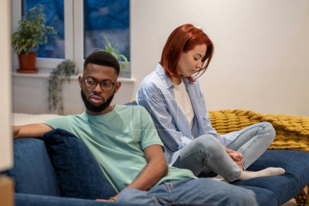 Photo for Unhappy young interracial couple sit separately on sofa at home, tired of each other, upset family man and woman not talking after fight, sit on couch apart. Bad communication in relationship - Royalty Free Image
