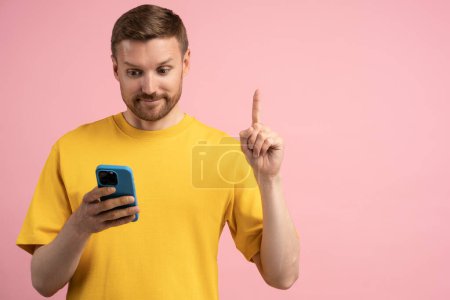 Foto de Hombre interesado muestra signo de dedo Idea recibir oferta por mensaje en el teléfono móvil en fondo de estudio rosa. Sonriente chico se para con gesto de atención extiende el dedo índice hacia arriba mirando en la pantalla del teléfono celular - Imagen libre de derechos