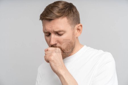 Photo for Sick man coughs due to illness at studio wall. Unhealthy male in white t-shirt with flu symptoms feels discomfort sore throat stand on studio grey wall. Entry of contagious infection into human body - Royalty Free Image