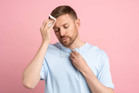 Photo for Man suffering from heat stuffiness wiping sweat on forehead with paper napkin isolated on pink background. Exhausted overheated bearded male with closed eyes. Summer hot weather, stuffy room concept. - Royalty Free Image