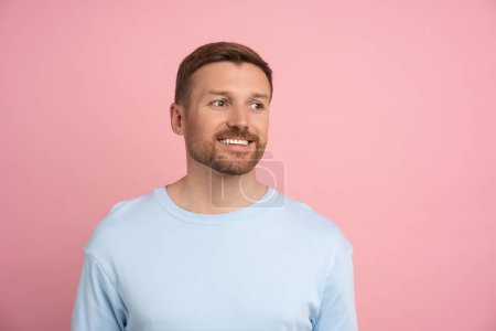 Téléchargez les photos : Homme positif avec sourire blanc regarde de côté sur fond rose. Concept de bonne humeur. Manifestation des émotions du gars joyeux pour la promotion de l'offre publicitaire dans le mur studio espace de copie vide - en image libre de droit