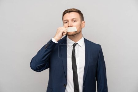 Photo for Confident man in formal suit with taped mouth tearing off tape from lips isolated on gray studio wall. Silent male entrepreneur not able to speak mind bravely removes restrictions at work. Censorship - Royalty Free Image