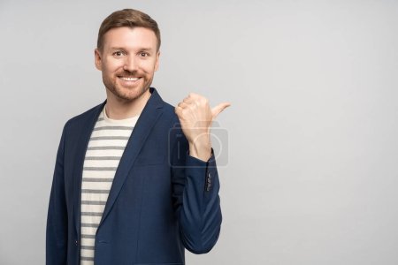 Photo for Happy friendly bearded man in jacket showing thumb finger on copy space on grey studio background through shoulder looking at camera. Advertisement banner, poster. Smiling guy businessman advertising. - Royalty Free Image