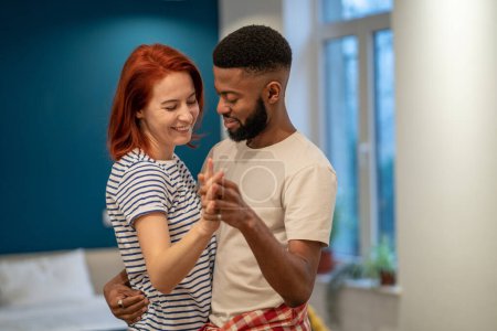 Téléchargez les photos : Happy aimant jeune couple diversifié apprenant à danser la danse romantique dans un appartement moderne. Souriant mari noir et femme allemande célébrant l'anniversaire, appréciant le moment tendre, s'amusant à la maison - en image libre de droit