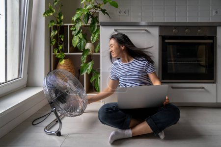 Photo for Woman turned powerful electric fan sitting cross-legged on floor in modern kitchen working on laptop cooling summer heat. Asian woman freelancer fighting effects global warming with floor ventilator. - Royalty Free Image