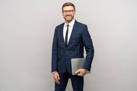 Photo for Portrait of handsome smiling businessman in suit, eyeglasses holds laptop on grey background looking at camera. Successful guy entrepreneur, office worker posing for advertisement banner, poster. - Royalty Free Image