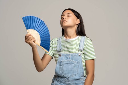 Photo for Overheated teenage girl holding bright fan in her hand. Tired teenager girl languishing from heat fanning herself trying to cool isolated on studio gray background. Health impact of global warming - Royalty Free Image