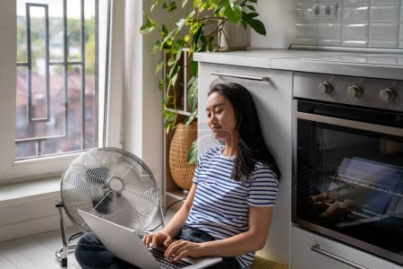 Photo for Relaxed Asian woman typing on laptop cooling by floor electric fan sits lotus position on floor in kitchen. Carefree Chinese girl blogger inspiredly writes article online magazine, work at home - Royalty Free Image