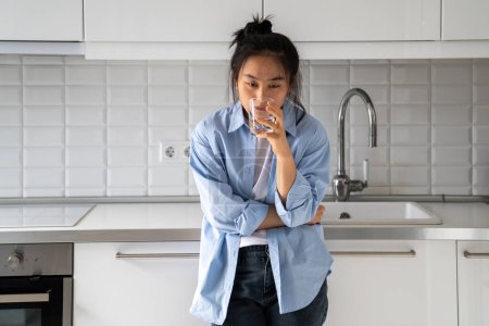 Photo for Unhappy disheveled Asian girl drinks water from glass while standing in kitchen waking up late morning bad mood, drinking an antidepressant. Korean middle-aged sick woman indigestion and thirsty. - Royalty Free Image