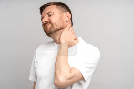 Photo for Man suffers from neck pain. Tormented by pain, guy holds his neck with hand with suffering expression on face. Protrusion, pinching, pinched nerve, sedentary work, hernia. Isolated on gray background. - Royalty Free Image