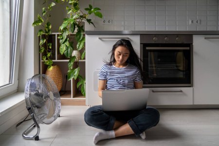 Photo for Focused asian girl student with laptop is languishing heat sitting on cool floor next fan and open window in modern apartment. Contented Chinese woman enjoying cool wind home at floor air conditioner. - Royalty Free Image