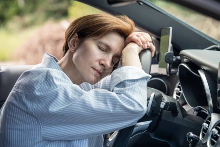 Foto de Mujer cansada de mediana edad que duerme en el volante agotado de la conducción larga en el viaje por carretera. Conductora siente fatiga descansando teniendo descanso en la carretera. Viajar en coche, viajar en larga distancia. - Imagen libre de derechos