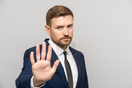 Man businessman gestures to stop. Young Caucasian bearded business guy in suit shows open palm meaning stop enough urging to avast. Fatigue from annoying office rules, protest, desire to change system