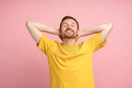 Photo for Well-rested smiling man on pink studio background. Carefree happy guy with hands behind neck in open relaxed pose smiles broadly, closed eyes. Contentment, happiness, satisfaction, euphoria, ecstasy. - Royalty Free Image