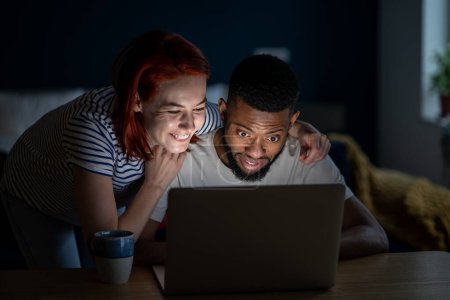 Téléchargez les photos : Heureux couple romantique regarder des vidéos sur Internet depuis un ordinateur portable. Homme et femme rient, sourient, s'émerveillent avec du contenu en ligne sur les réseaux sociaux, apprécient le film comique drôle. Mariage interethnique, épouse, mari - en image libre de droit