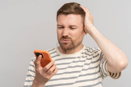 Photo for Awkward man looking at phone screen. Embarrassing, difficult, clumsy, uncomfortable message, news in social networks on Internet online. Guy wrinkled with embarrassment, shame looks at smartphone - Royalty Free Image