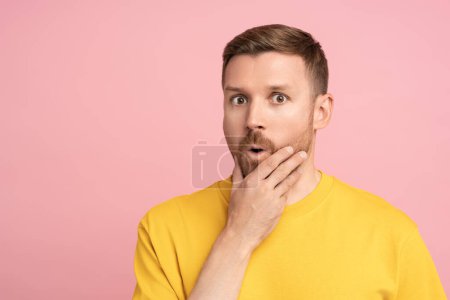 Photo for Surprised man holds on to chin. Astonished, interested brown haired young guy listens carefully to gossip, rumors, stories with mouth open. Feeling of amazement wonderment. Isolated on studio pink. - Royalty Free Image
