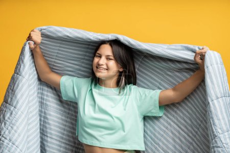 Photo for Smiling teenage girl in blanket isolated on studio yellow wall. Happy joyful satisfied pleased cheerful girl enjoys comfort, coziness in warm soft white gray duvet created for long sound healthy sleep - Royalty Free Image