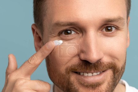 Photo for Pleased handsome man applying skincare lotion product on face. Attractive male with perfect skin put cosmetic cream smiling against blue background. Lifting moisturising anti-aging procedure for guy - Royalty Free Image