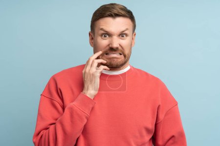 Ekelgefühle im Gesicht des jungen Mannes. Mann mittleren Alters in rotem Sweatshirt auf blauem Hintergrund mit hellem, ausdrucksstarkem Gesichtsausdruck blickt mit Feindseligkeit und Abscheu in die Kamera.