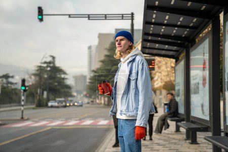 Photo for Young student guy waiting for bus. Man gen Z hipster with coffee cup relaxed awaits at bus stop station for trolleybus minibus to university college educational institution. Public urban transport use - Royalty Free Image