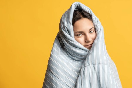 Photo for Smiling teenage girl wrapped in warm blanket peeking out with sleepy eyes isolated on studio yellow background. Morning winter mood, time go school. Peaceful woman lazily waking up for work - Royalty Free Image