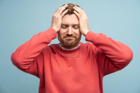 Photo for Irritated man with closed eyes holds head feeling discomfort due to headache on blue wall background. Concept of illness and suffering from unpleasant sensations. Sad bearded male regrets a mistake - Royalty Free Image