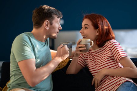 Photo for Happy man woman drinking tea talking smiling sitting on couch at home. Positive loving couple flirting enjoying time together. Young family everyday life, good romantic relationship love care concept. - Royalty Free Image