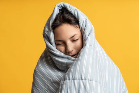 Téléchargez les photos : Portrait de femme brune endormie enveloppée dans une couverture rayée grise sur fond jaune. Fille avec les yeux fermés couverts de couette chaude. Confort confortable, manque de sommeil, sieste, insomnie, troubles du sommeil. - en image libre de droit