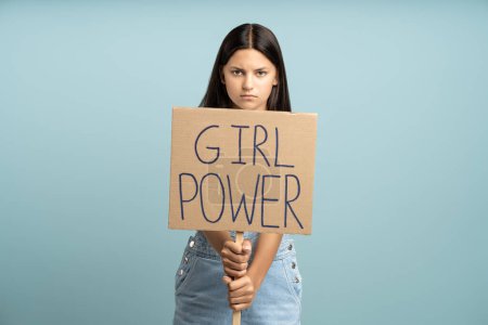 Photo for Serious confident brunette teenage girl showing holding girl power poster on blue background looking at camera. Fight of women rights, against harassment, violence, for feminism, demonstration concept - Royalty Free Image