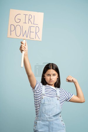 Photo for Confident strong brunette teenager girl with long hair showing power flexing arm holding girl power poster on blue background looks at camera. Woman independence, gesture with clenching fist concept. - Royalty Free Image
