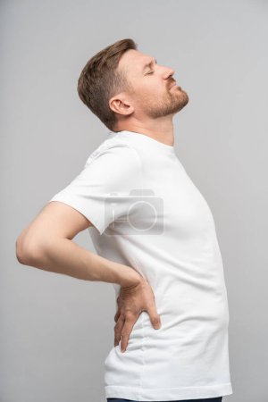 Photo for Backache from sedentary lifestyle. Frowning tired man touch small of back feels acute pain isolated studio background. Office syndrome passive life. Male with closed eyes writhing in sick discomfort - Royalty Free Image