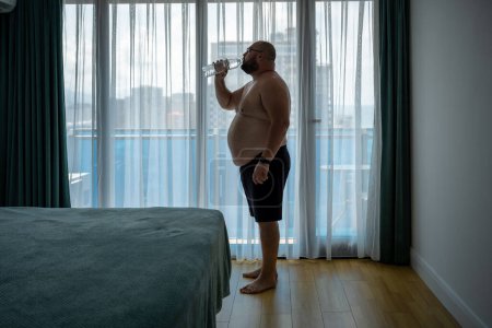 Photo for Thirsty overweight man feeling dehydration drinking water in hotel room in hot humid weather standing near window. Fat guy with naked torso suffers from heat, heat stroke, overheating, excess weight. - Royalty Free Image