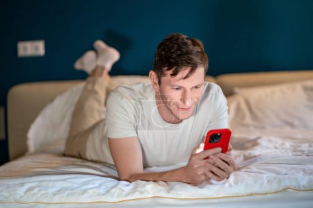 Photo for Man reading information in smartphone lying in bed at home in modern bedroom in evening. Interested concentrated male searching info, surfing online, watching content, scrolling social media in phone. - Royalty Free Image