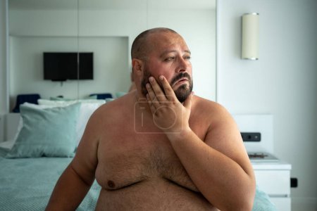 Photo for Portrait of overweight naked bearded man in air-conditioned room escaping from heat in hotel sitting in bed. Obesity male, health problems, plump chubby guy suffer from hot, stuffiness in summer. - Royalty Free Image